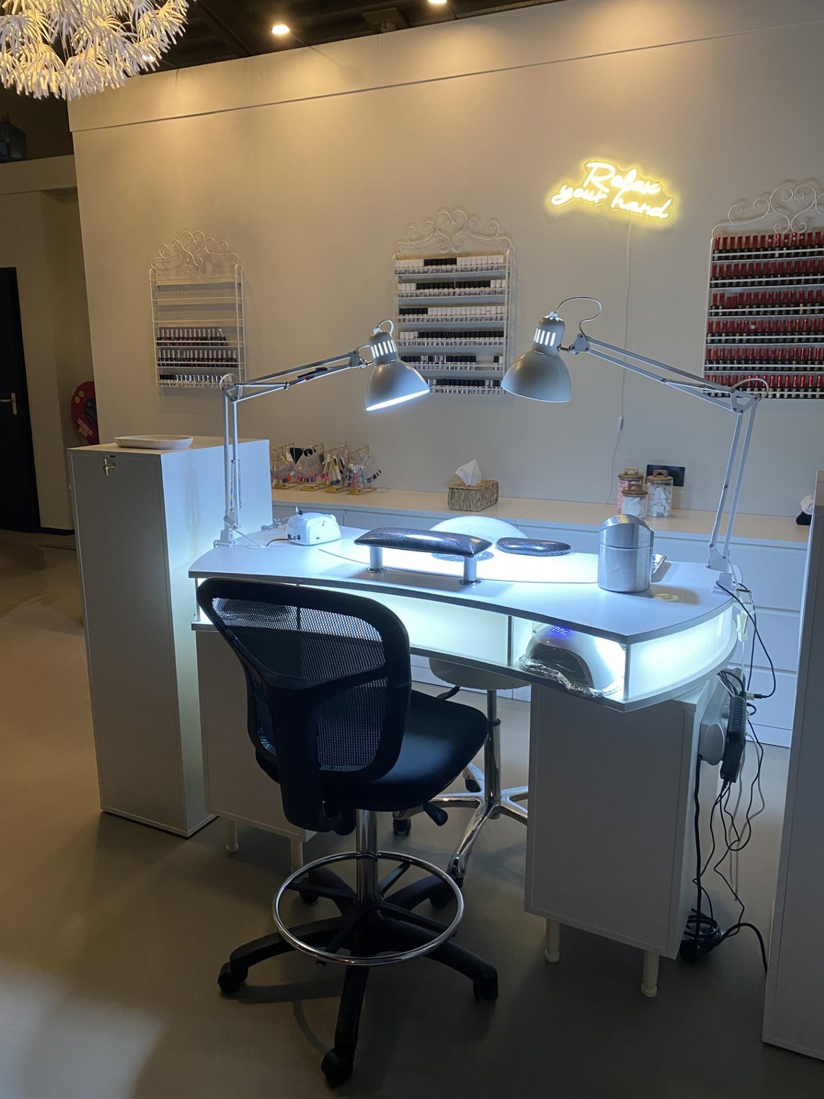 De witte nageltafel met twee lampen, een rek met lakjes en een neonbord aan de wand